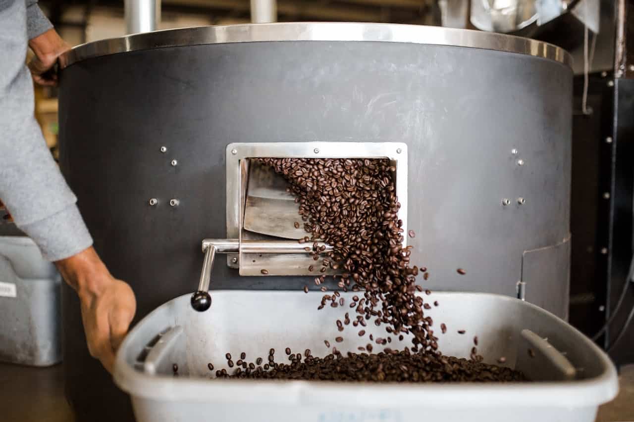 Coffee beans coming out of coffee roaster into a large plastic pan. A person's hand steadies the pan.